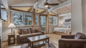 Amplia sala de estar con techo de madera, ventanas grandes con persianas, un sofá, una mesa de centro y una cama con colcha blanca.