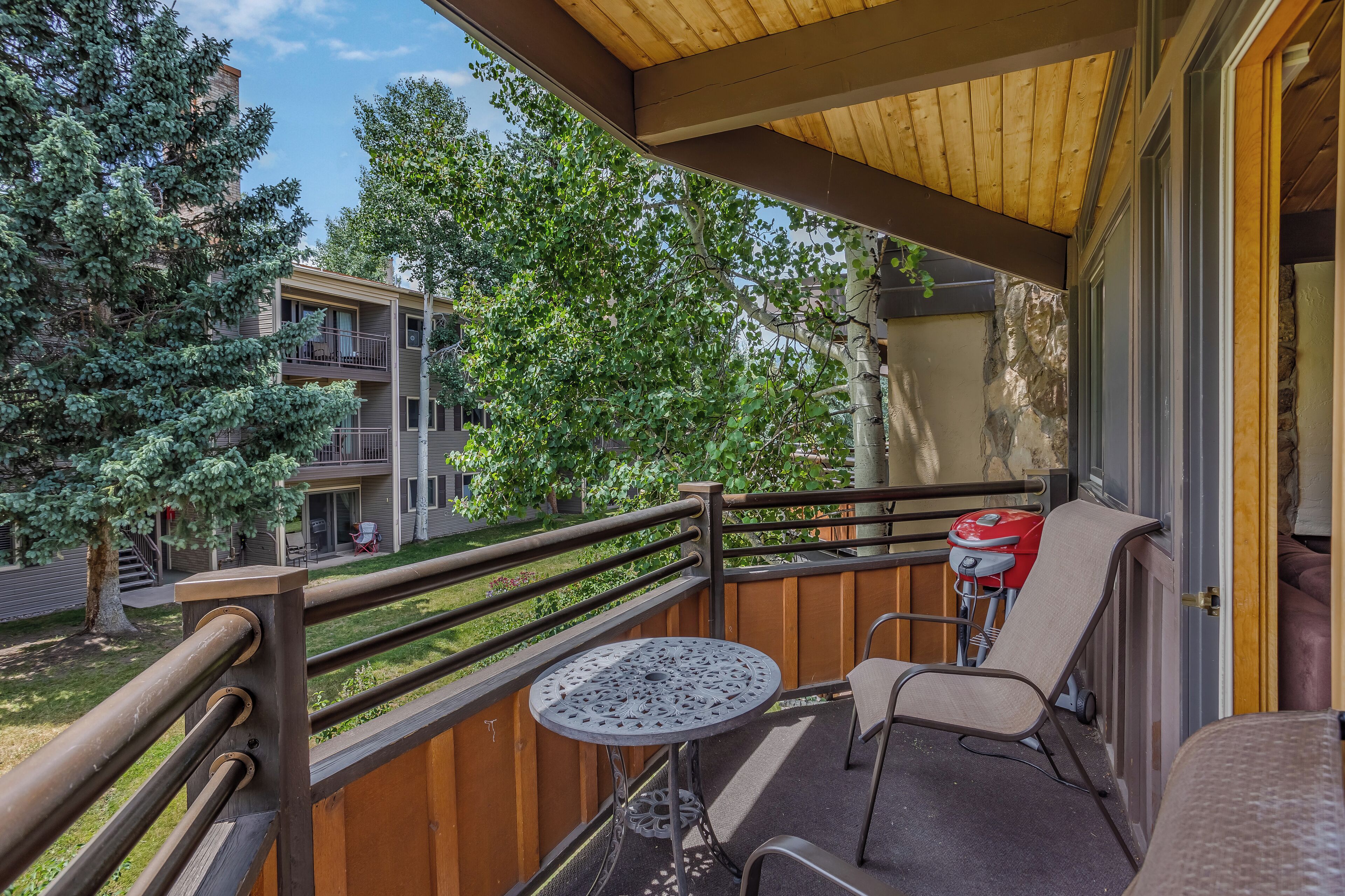 A balcony with a table and chairs, a wooden railing, and a view of neighboring buildings and trees.