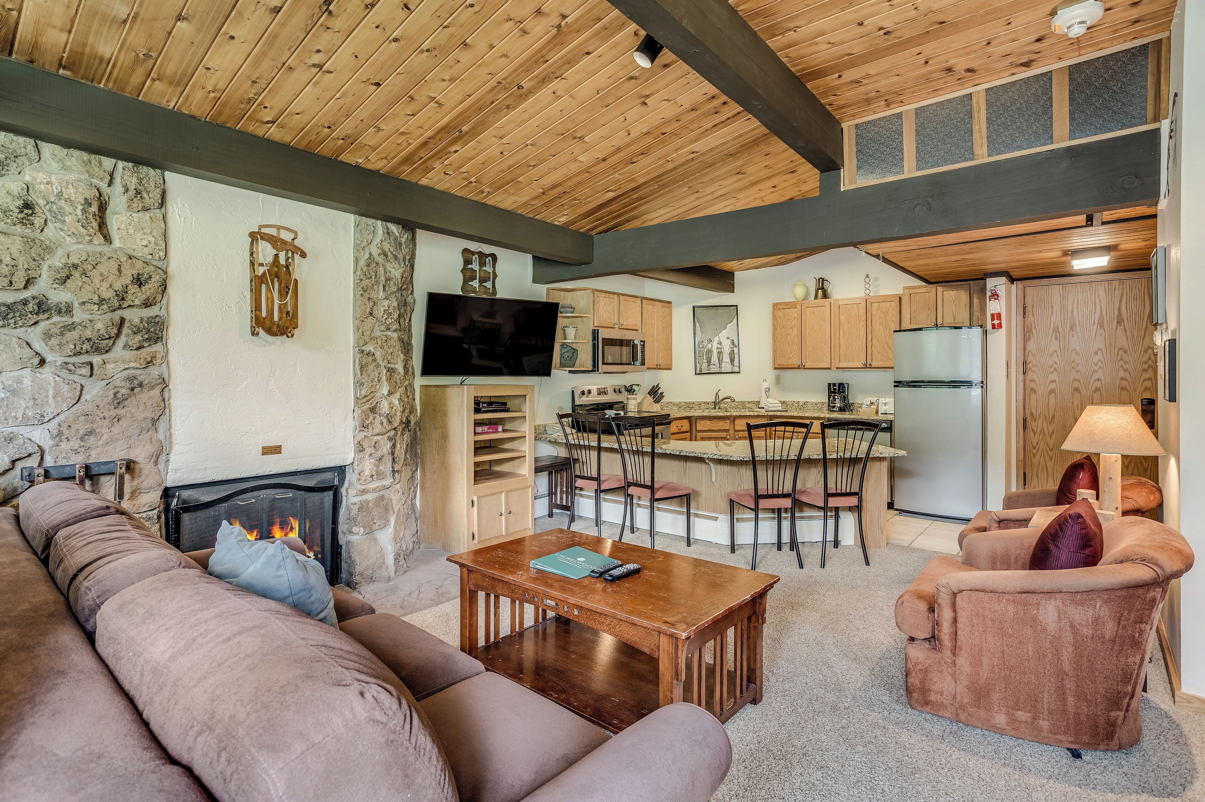 A cozy living room with a fireplace, a stone wall, and a kitchen in the background.