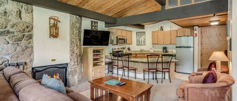 A cozy living room with a fireplace, a stone wall, and a kitchen in the background.