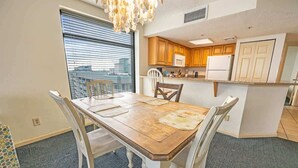 A dining area with a wooden table and white chairs, a kitchen with wooden cabinets and white appliances, and a large window with blinds. - Contemporary 3BR Beach Colony Resort -  Oceanfront (Myrtle Beach)