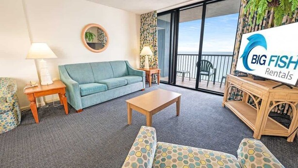 A living room with a sofa, coffee table, and patterned chairs. A sign for Big Fish Rentals is visible. - Contemporary 3BR Beach Colony Resort -  Oceanfront (Myrtle Beach)