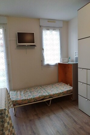 A small, single-room apartment with a bed, a TV mounted on the wall, a window with curtains, and a kitchenette area. - Les Minimes LA Rochelle Charente Maritime (La Rochelle)