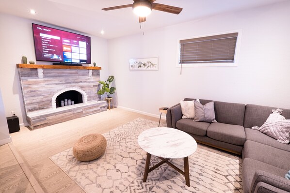 Una sala de estar con chimenea de piedra, un televisor de pantalla plana montado encima, un sofá gris, una mesa redonda y una alfombra estampada.