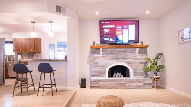 A modern living room with a stone fireplace, a flat-screen TV mounted above it, and a seating area with two bar stools. - 🌵 Cozy Cactus Home near Old Town & ASU 🌵 Pool ⭐ Game Room ⭐ BBQ ⭐ Fire Pit ⭐ (Tempe)