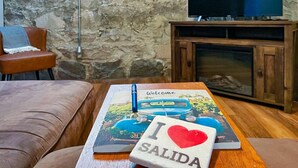 A cozy living room with a stone wall, a television on a wooden stand, a leather sofa, and a table with a book and a card. - The perfect spot in Salida - F Street Hideaway! (Salida)