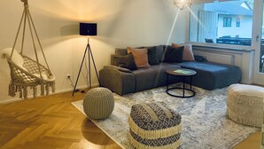 A cozy living room with a sofa, hanging chair, floor lamp, ottoman, and a view of a building outside. - Modern vacation apartment "La Fredo" near Lake Starnberg, 5 minutes' walk to public transport (Wolfratshausen)