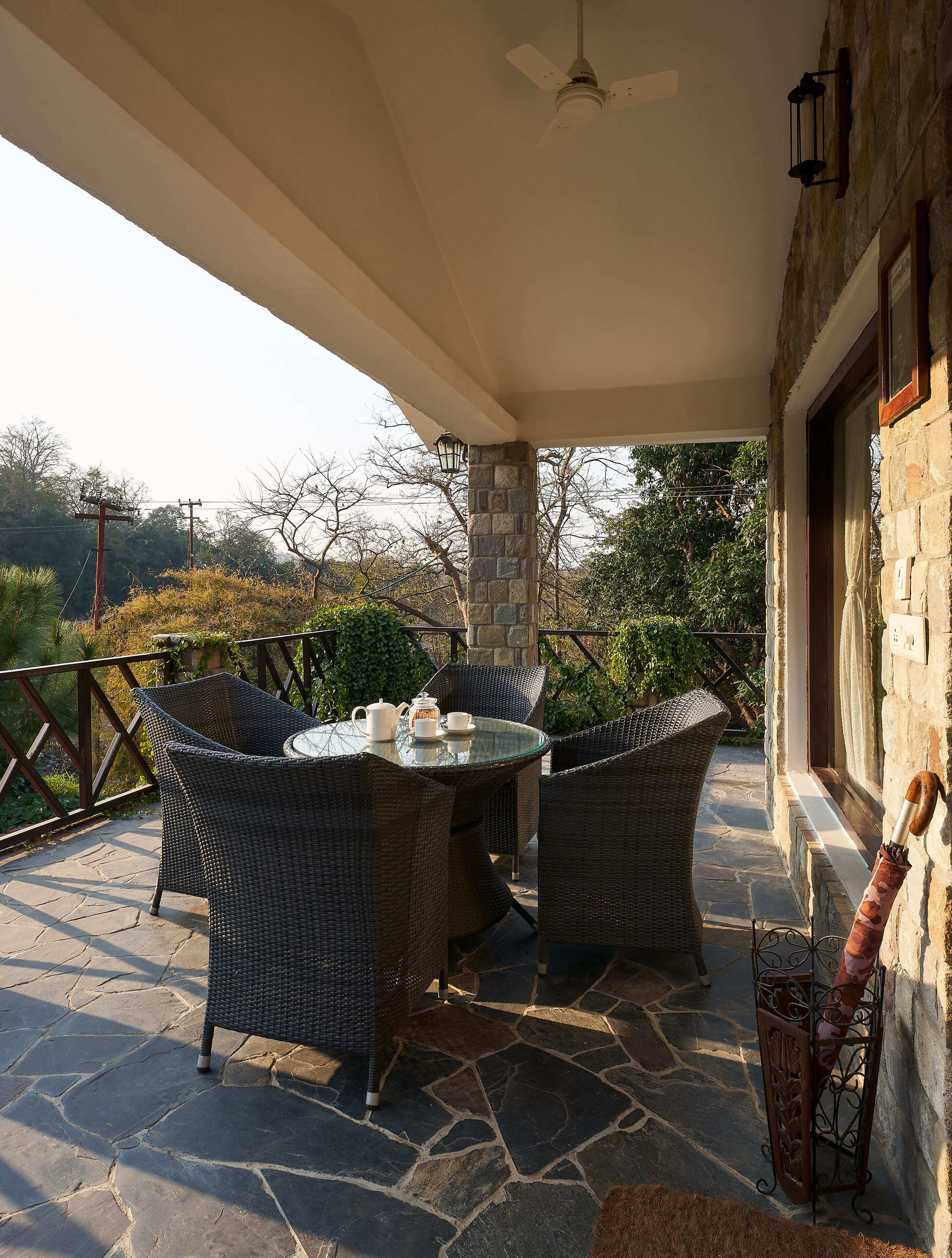 A covered patio with wicker furniture, a table with a teapot and cups, and a stone floor.