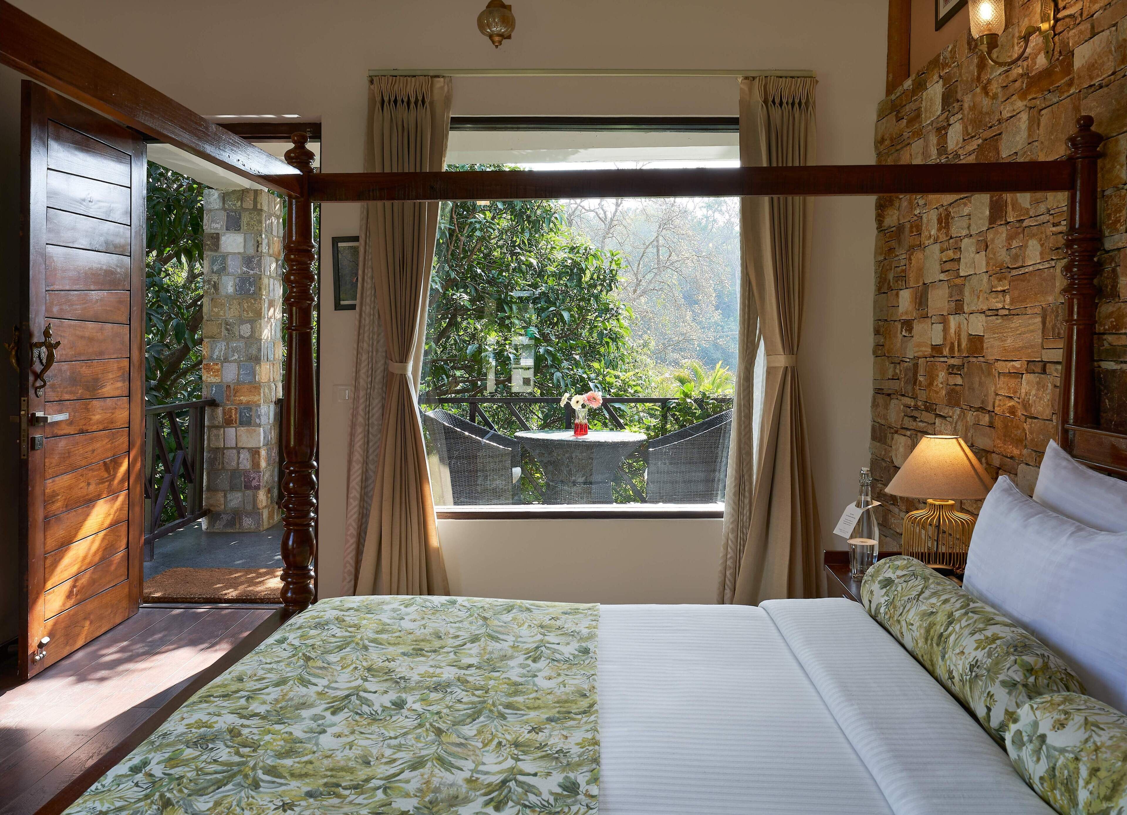 A bedroom with a large bed, a stone wall, a window with a view of greenery, and a wooden door.