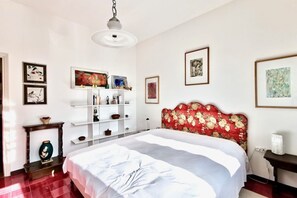 A bedroom with a bed, a white wall with framed artwork, a wooden side table, and a red floral headboard. - General Terms and Conditions - Vacation Rental:Welcome to our vacation rental. To ensure a pleasant and hassle-free stay, please review the following general terms and conditions.Assistance ServicesIn-person assistance: Available from 9:00 AM to 6:00 PMT (Dervio)