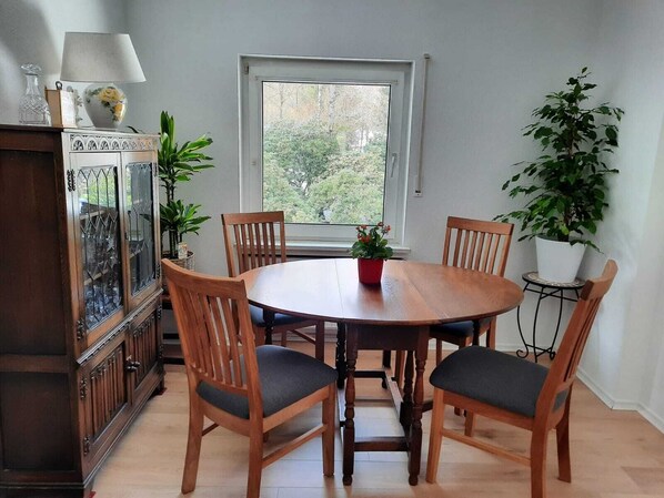 Un comedor con una mesa redonda de madera, cuatro sillas de madera con asientos acolchados, un aparador con puertas de vidrio, una ventana con vista a árboles y una planta en maceta en el alféizar.