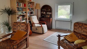 Un salón acogedor con una estantería de madera, un sillón cómodo, un sofá estampado y una ventana con vistas al verdor.