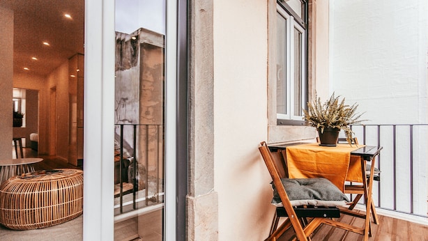Ein Balkon mit einem Korbsitzhocker, einem Holzstuhl mit Kissen und einem Blumentopf.