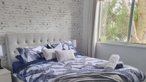 A bedroom with a brick-patterned wall, a tufted headboard, a bed with a patterned duvet, a bedside lamp, and a window with a view of trees. - The Sandbar (Lake Conjola)