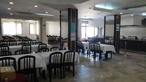 Un grand espace dédié à la salle à manger, avec plusieurs tables et chaises, un coin cuisine à l’arrière-plan et une vue dégagée sur l’entrée.