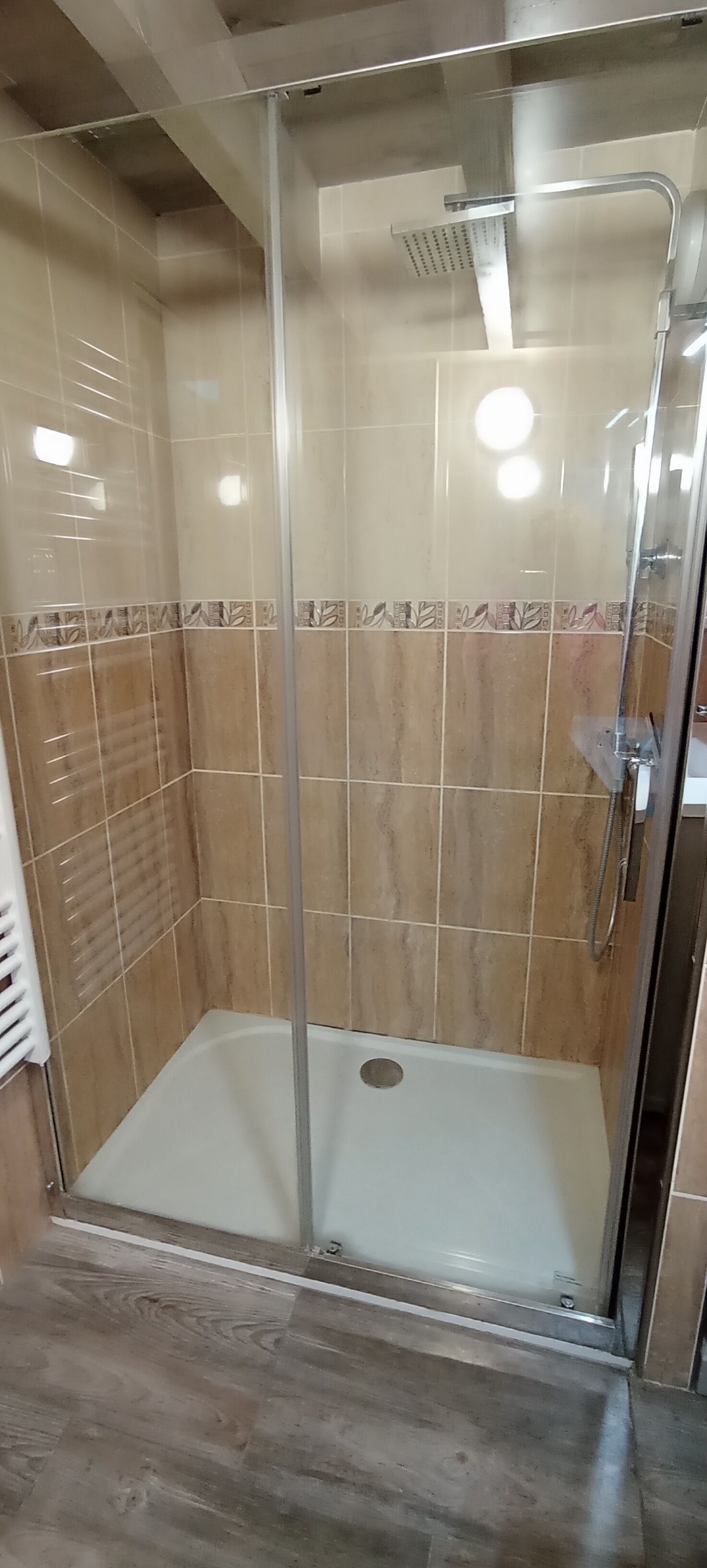 A glass-enclosed shower with a white bathtub, tiled walls, and a wooden floor.
