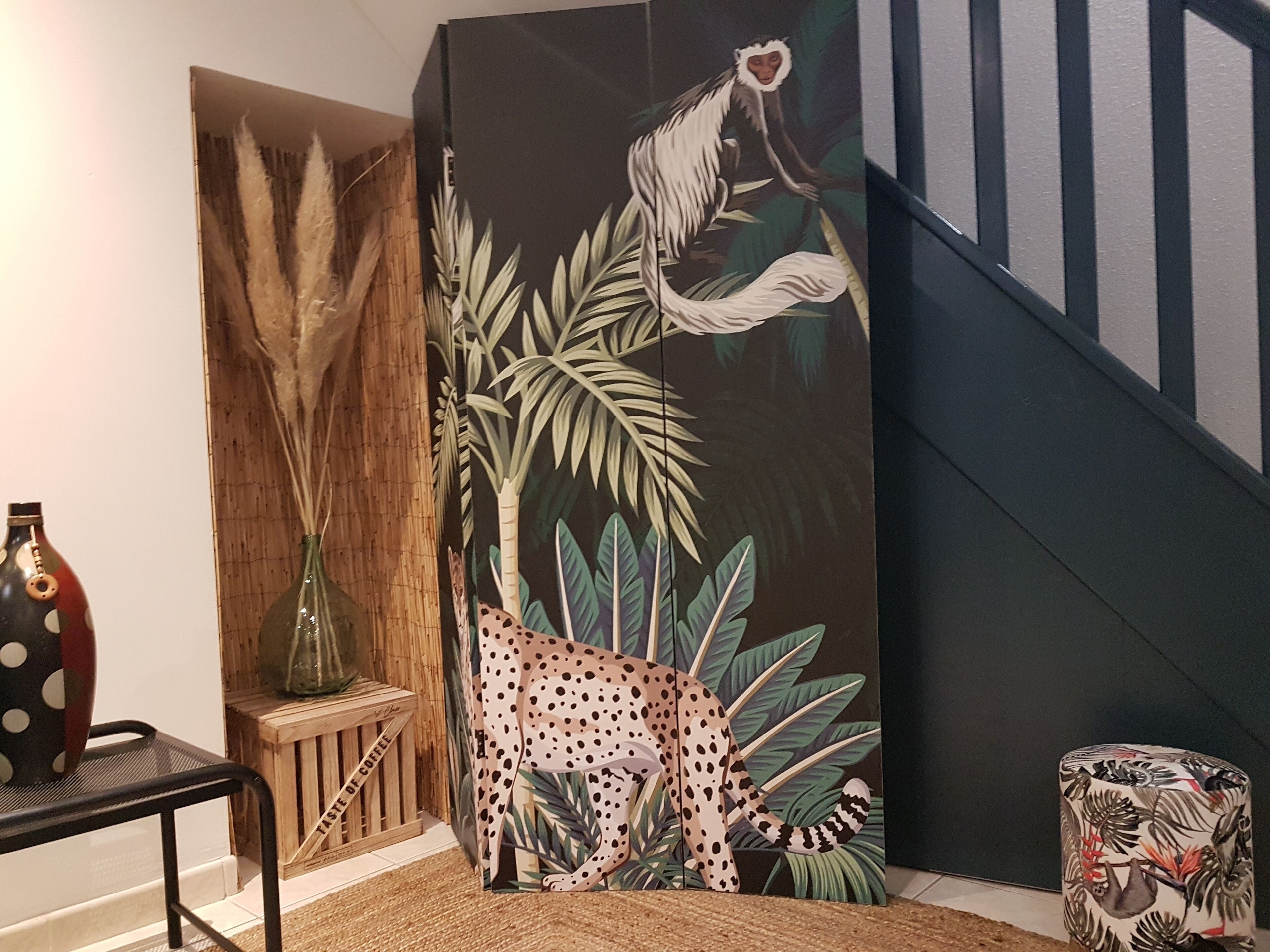 Une pièce décorée d’un grand tableau aux motifs tropicaux, d’une caisse en bois étiquetée « GOÛT DE CAFÉ » et d’un pouf à motifs.