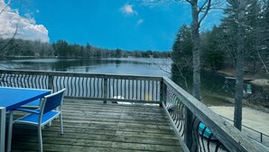 A wooden deck with a table and chairs, overlooking a calm lake with trees in the background. - Waterfront home on Perch Lake outside Traverse City. Hot tub. Pool table. (South Boardman)