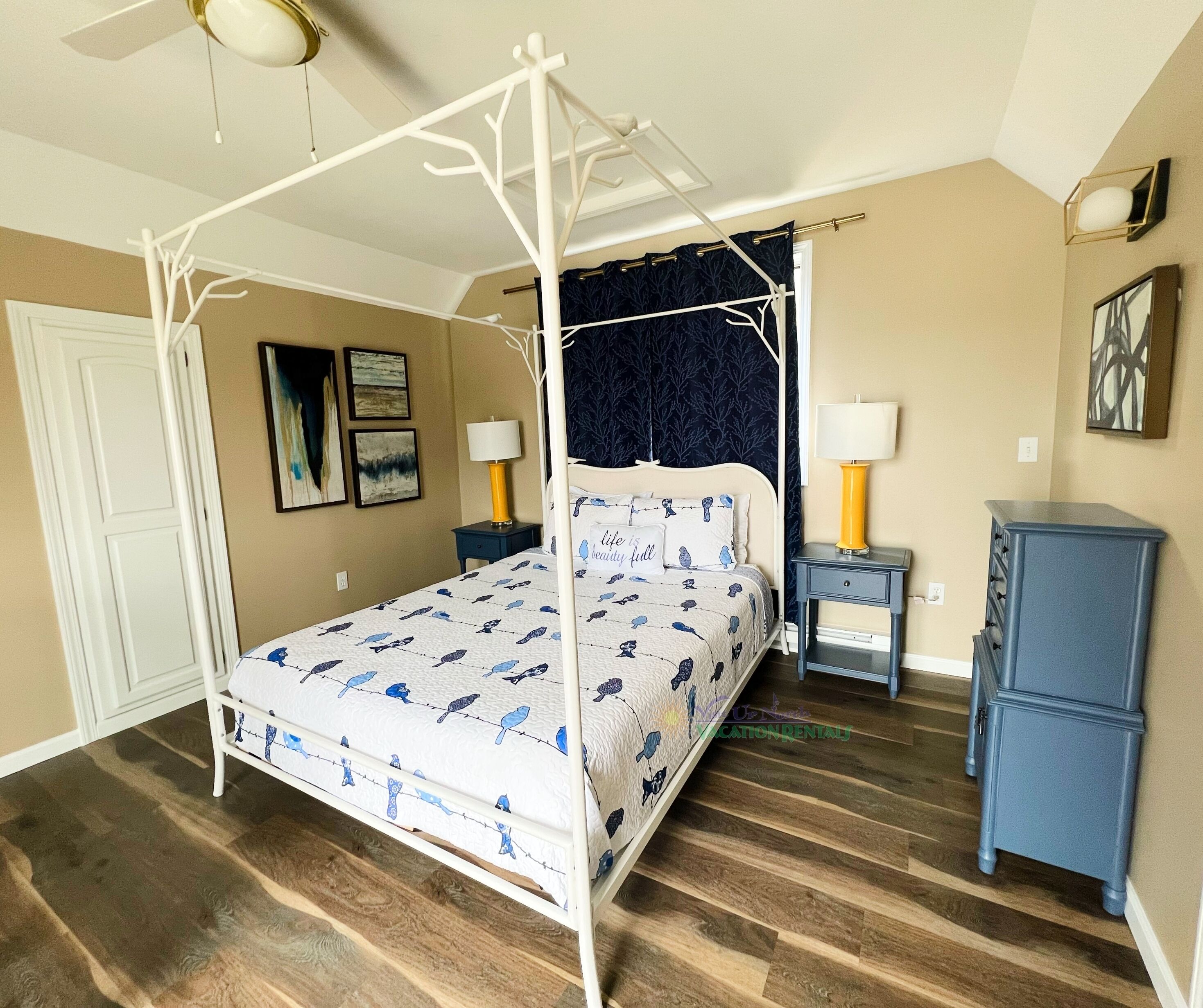 A four-poster bed with a white frame, a dark blue canopy, and a patterned bedspread. There are two bedside tables with lamps, a dresser, and a door to the left.
