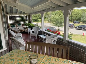 A covered porch with wicker furniture, a table with a floral tablecloth, and a view of a parking lot and trees.