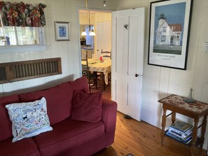 A cozy living room with a red sofa, a wooden bench, and a small table with books. A door leads to a kitchen area with a dining table and chairs.