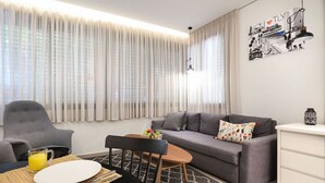 Ein modernes Wohnzimmer mit einem grauen Sofa, einem kleinen Tisch und einem Sessel. Ein weißes Schränkchen und ein Bild sind an der Wand zu sehen.