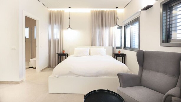 Ein modernes Schlafzimmer mit einem großen Bett, einem grauen Sessel und einem Fenster mit Jalousien.