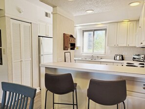 Cocina con gabinetes blancos, un refrigerador, un lavamanos y dos taburetes de bar.