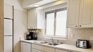 A kitchen with white cabinets, a dishwasher, a coffee maker, a toaster, and a window with blinds. - Townhouse Style Condo, Beach Access Across Road, Pool, Grilling Area, Elevator (Pawleys Island)