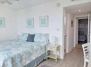 A bedroom with a bed, bedside lamps, a nightstand, and a door leading to another room. - Fourth Floor Condo across from Beach Access, Balcony, Pool, Grill Area, Elevator (Pawleys Island)
