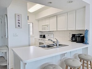 A modern kitchen with white cabinetry, a built-in oven, a microwave, a coffee maker, and a sink. - Fourth Floor Condo across from Beach Access, Balcony, Pool, Grill Area, Elevator (Pawleys Island)