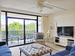 A spacious living room with a large TV, comfortable seating, a balcony with outdoor furniture, and a ceiling fan.