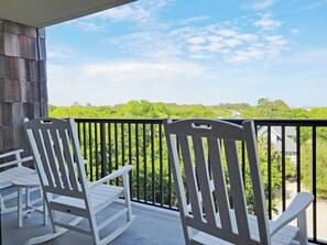 A balcony with white rocking chairs and a view of a green landscape. - Fifth Floor Condo across from Beach Access, Balcony, Pool, Grill Area, Elevator (Pawleys Island)