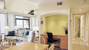 A room with a dining table, chairs, a sofa with floral upholstery, a desk with a chair, and a view of the outside through a window. - Top Floor Condo, Beach Access across Road, Balcony, Pool, Grill Area, Elevator (Pawleys Island)
