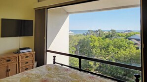 Une chambre à coucher comprenant un lit, une commode, un téléviseur à écran plat et une grande fenêtre donnant sur la mer et la verdure.