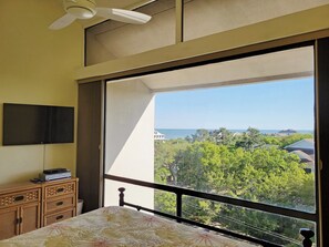 A bedroom with a bed, a dresser, a flat-screen TV, and a large window with a view of the sea and greenery. - Top Floor Condo, Beach Access across Road, Balcony, Pool, Grill Area, Elevator (Pawleys Island)
