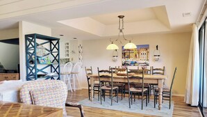 A dining area with a wooden table and chairs, a kitchen in the background, and a large window with curtains.