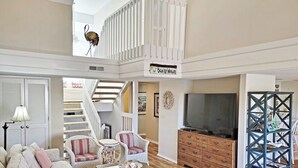 A spacious living room with a high ceiling, a staircase, a television, and a bookshelf.