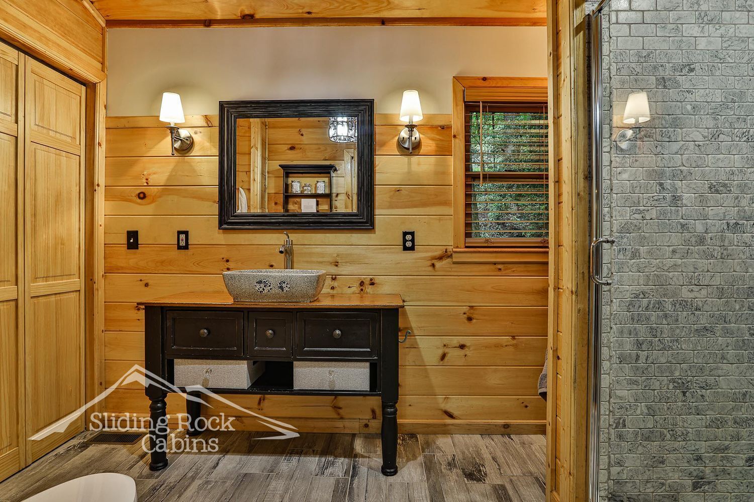 A bathroom with wooden paneling, a stone sink, two wall-mounted lights, a mirror, and a window with blinds.