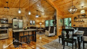 A cabin interior with a kitchen, dining area, and living space, featuring wooden walls and floors, and a stone fireplace.