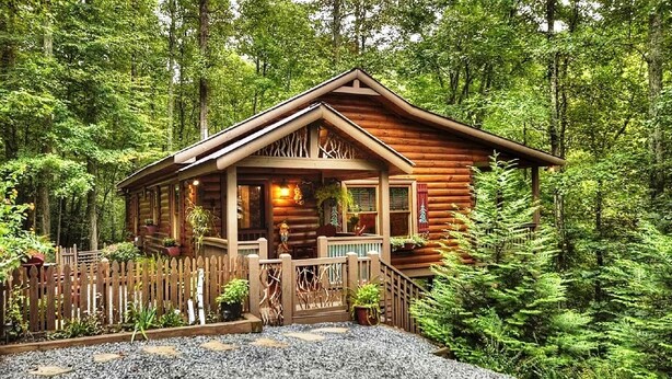 A log cabin with a porch, surrounded by a wooden fence and gravel path, set in a forest.