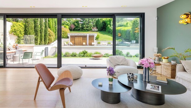 Un salon moderne doté d’une grande porte-fenêtre coulissante donnant sur un jardin. On y trouve un canapé, une table basse et un fauteuil.