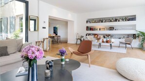 Un salon moderne avec un canapé, une table basse et des objets décoratifs. On y trouve également un coin repas avec des chaises et une table. La pièce est dotée de grandes fenêtres et d’une bibliothèque contenant divers objets.