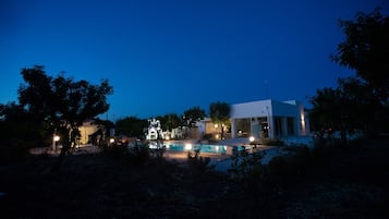 Une maison moderne avec une piscine, entourée d’arbres et illuminée par des éclairages extérieurs.