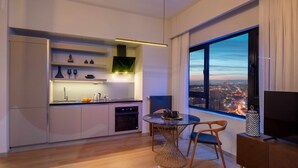 A modern kitchen with a dining area, large windows with a city view, and a TV on a wooden cabinet. - G Tower Residences (Istanbul)