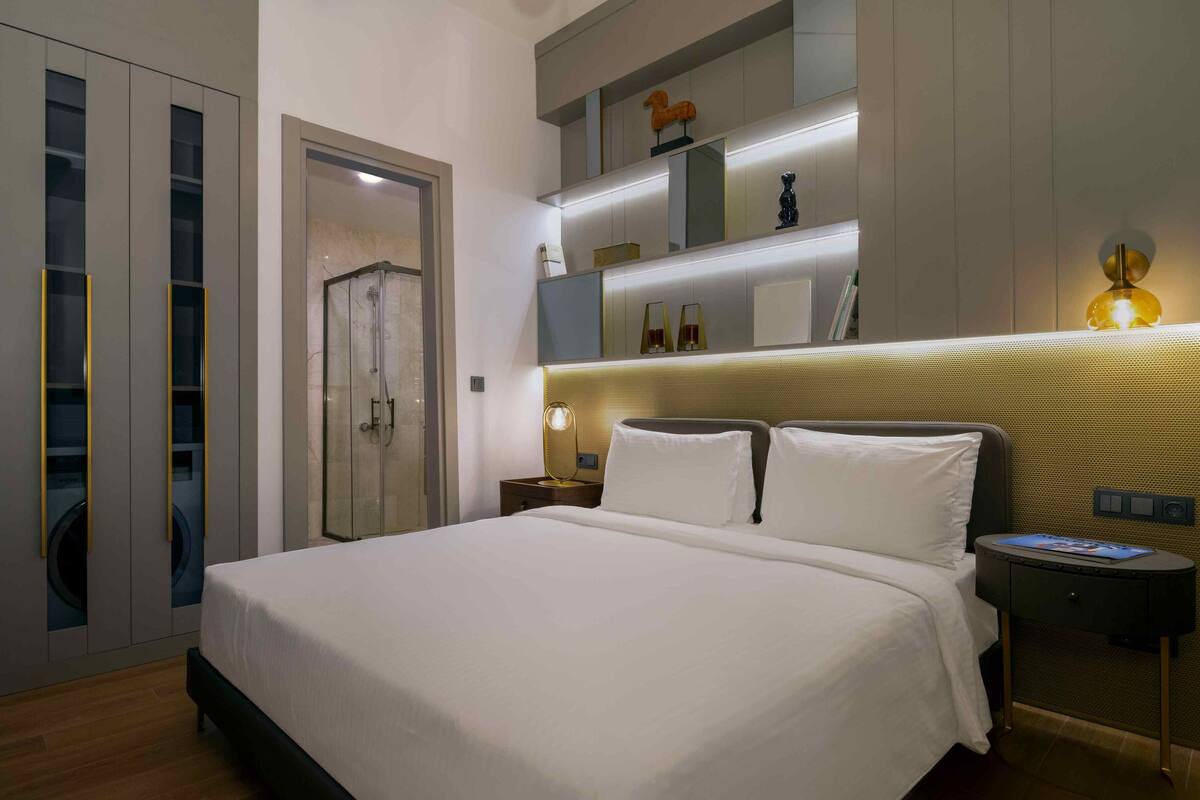 A modern hotel room with a large bed, a walk-in closet, and a headboard with built-in lighting.