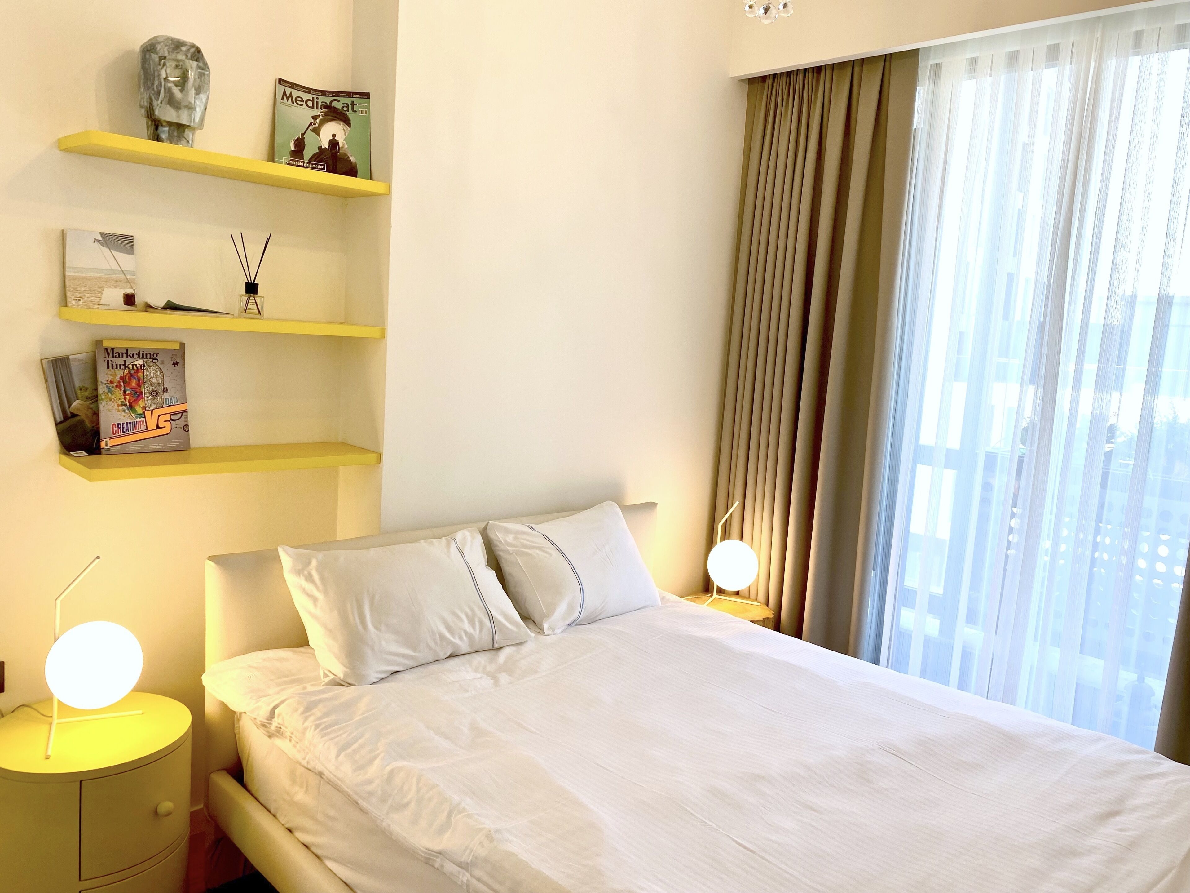 A hotel room with a bed, two bedside tables, a shelf with books and a lamp, and a window with curtains.