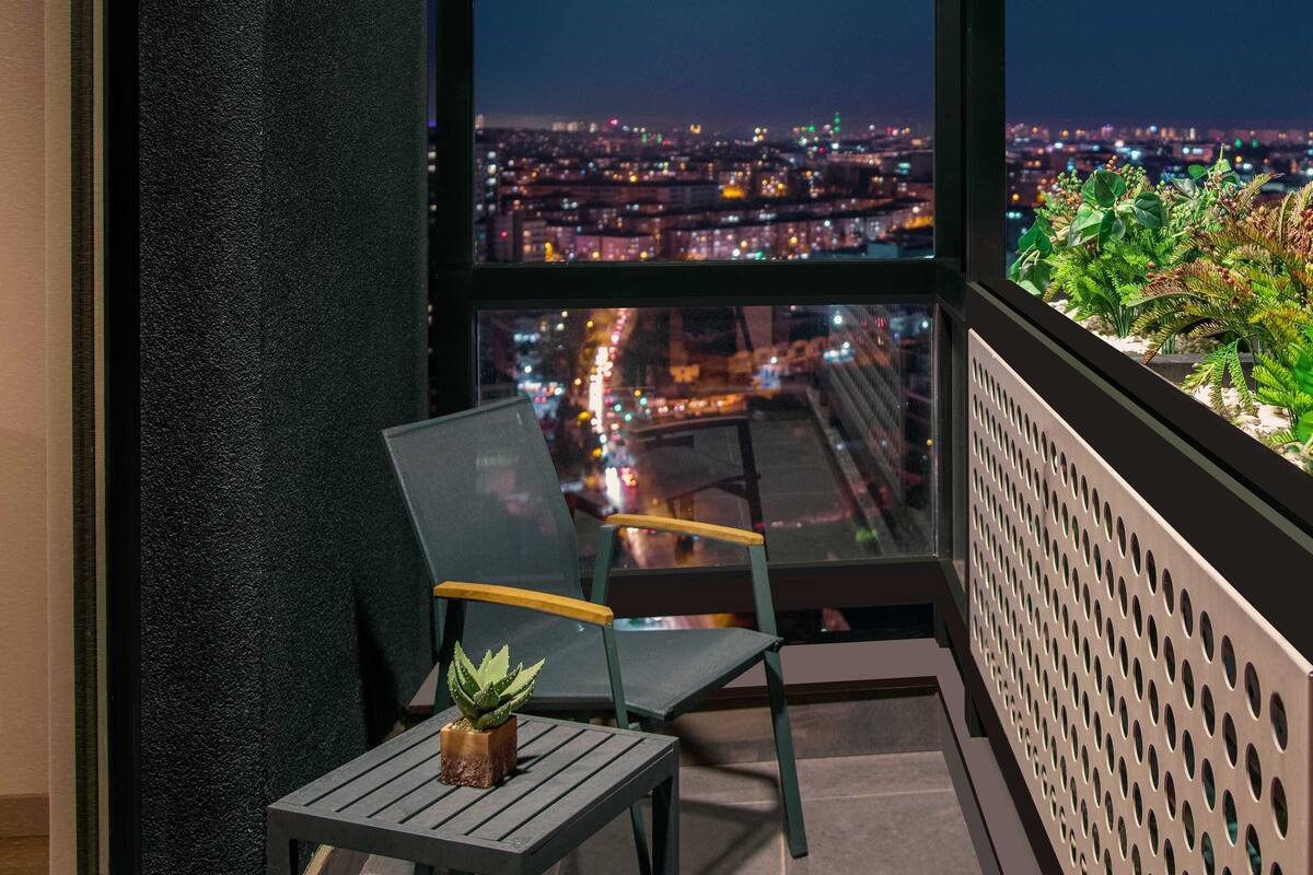 A balcony with a chair and a small table, a potted plant, and a city view at night.