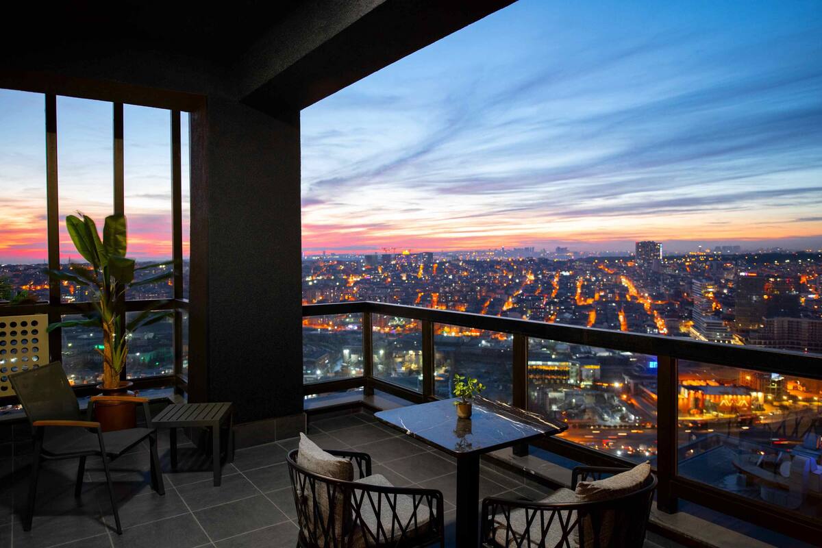 A balcony with a city view at dusk, featuring a table and chairs, and potted plants.