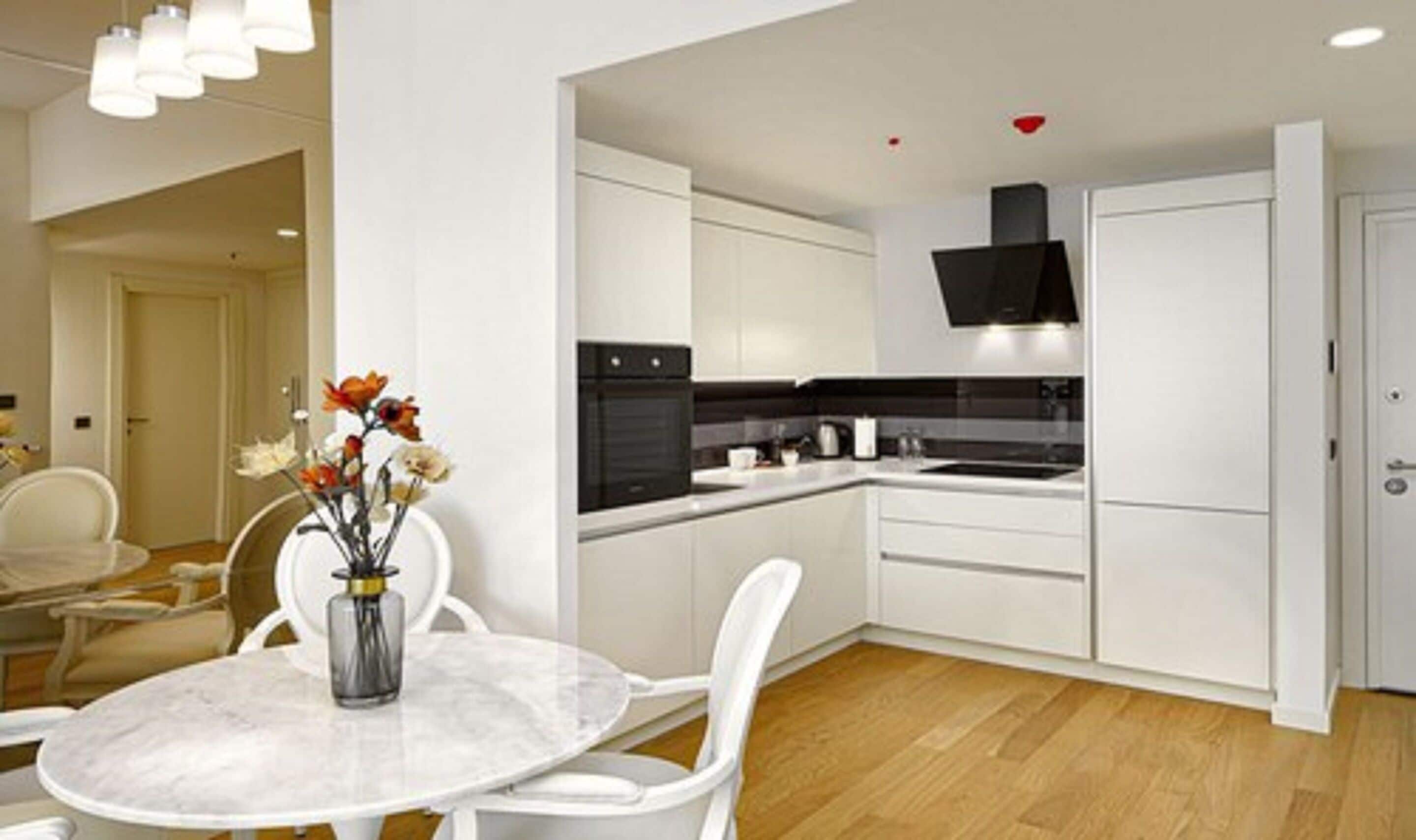 Una cocina moderna con armarios blancos, un horno negro y un microondas integrado. Hay una mesa de comedor con sillas blancas y un jarrón con flores.